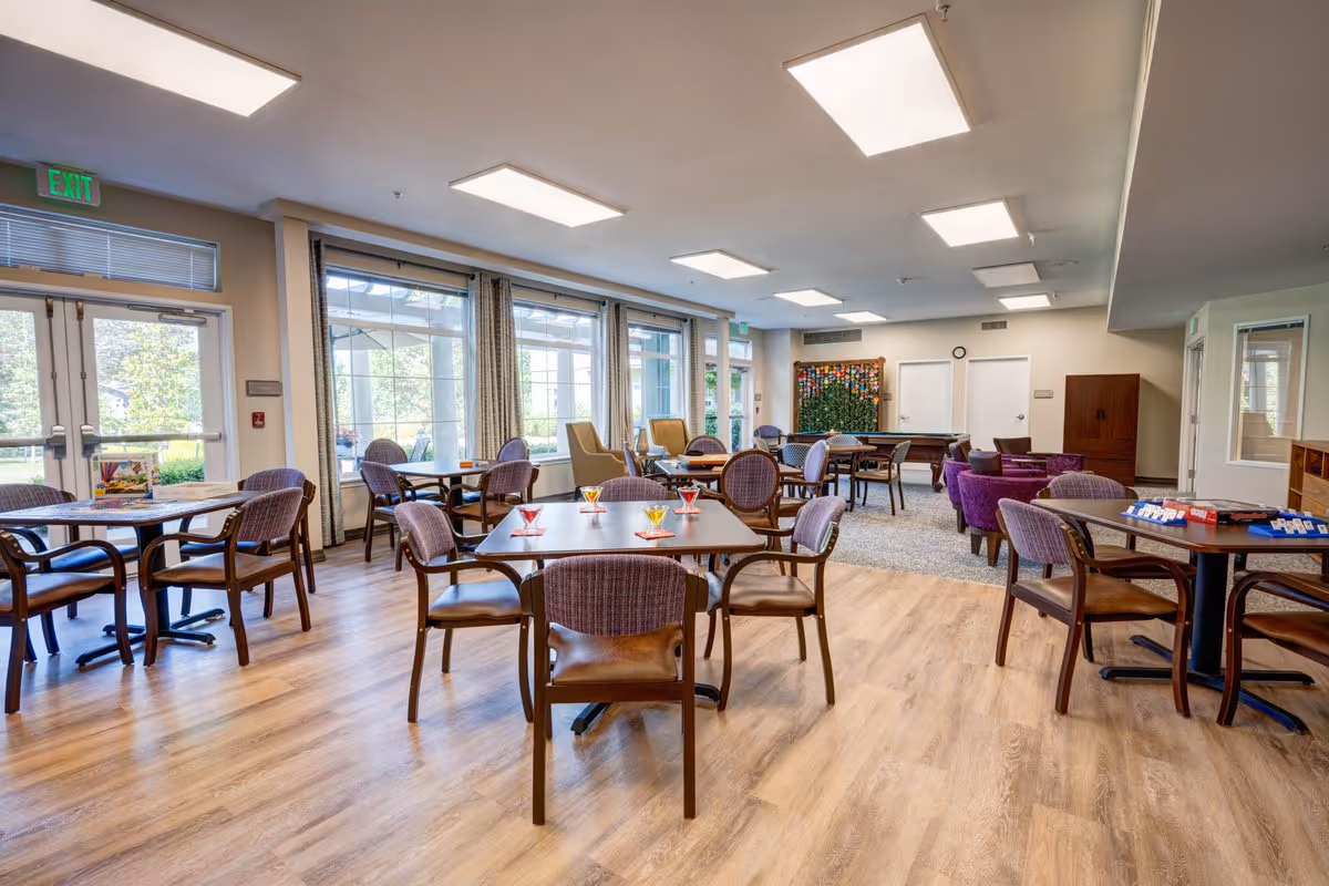 A spacious and well-lit common area in a senior living facility with multiple tables and chairs arranged for socializing or activities. Large windows with curtains allow natural light to fill the room. There are board games on some tables and comfortable armchairs in the background near a pool table and a decorative wall piece.