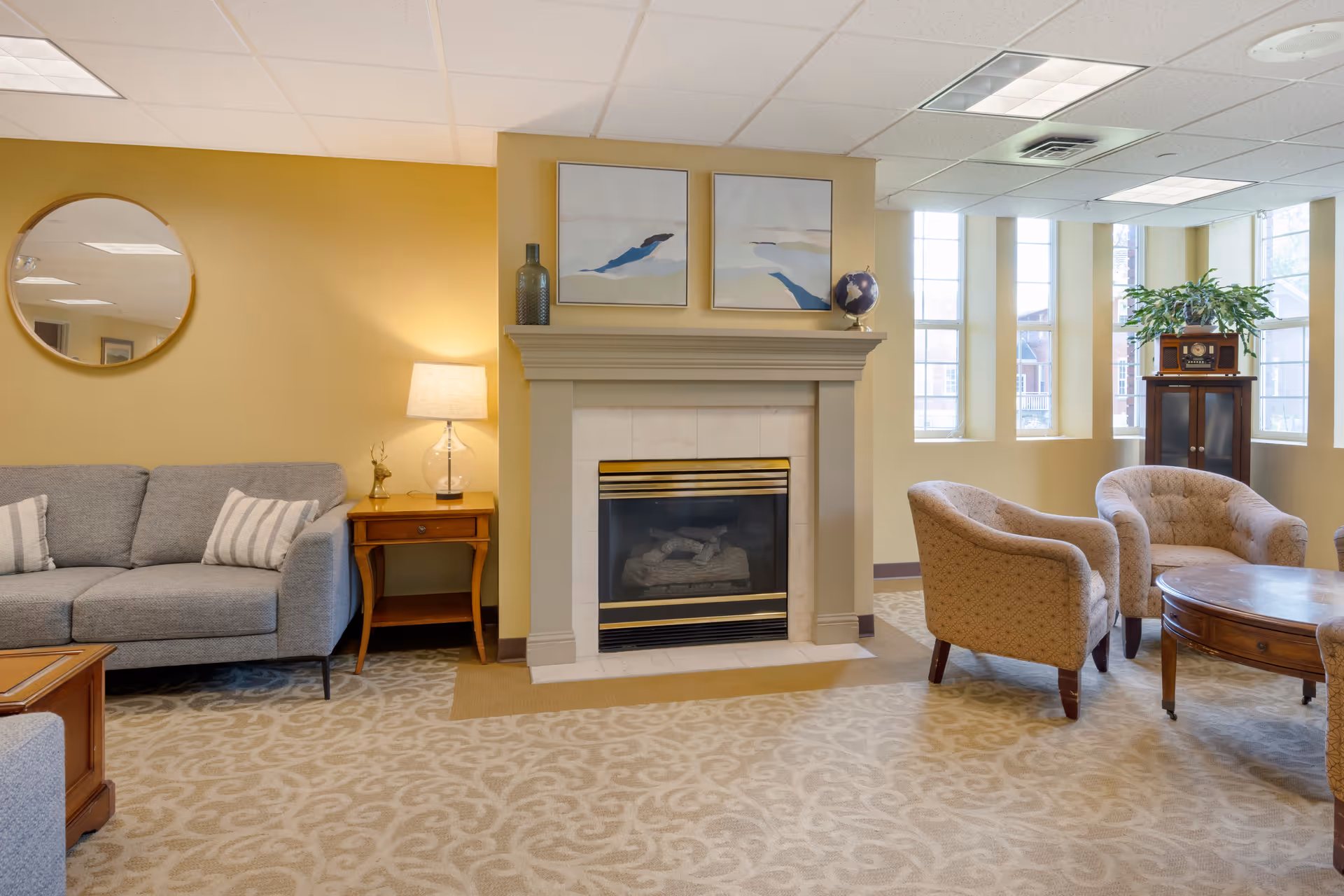 A cozy living room area with a gray sofa on the left, a wooden side table with a lamp, a fireplace in the center with two abstract paintings and decorative items on the mantel, two beige upholstered armchairs, a round wooden coffee table, large windows letting in natural light, and a wooden cabinet with a vintage radio and a plant on top.