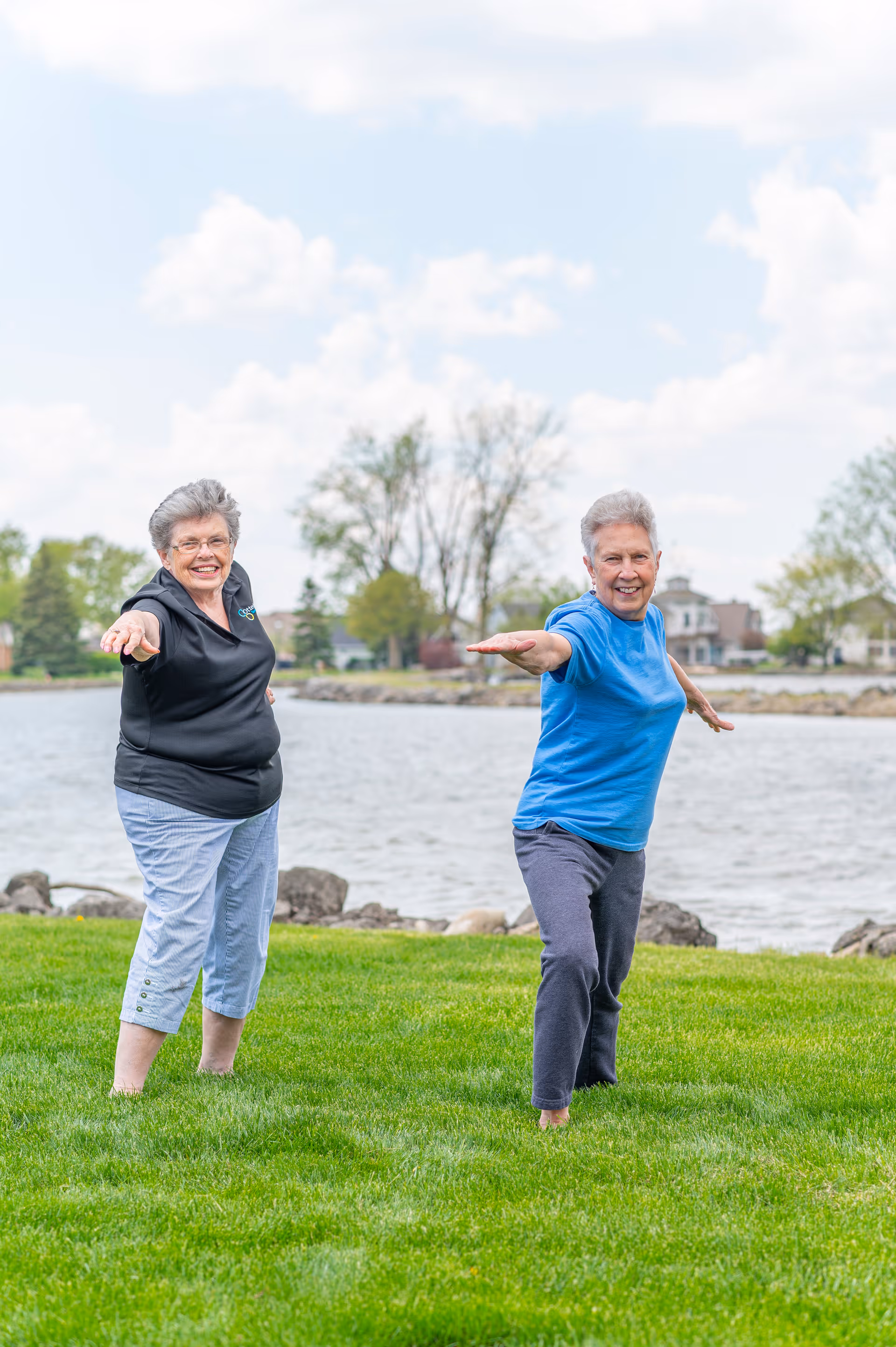 Two elderly women practicing yoga or stretching exercises on a grassy area near a body of water with trees and houses in the background under a partly cloudy sky.