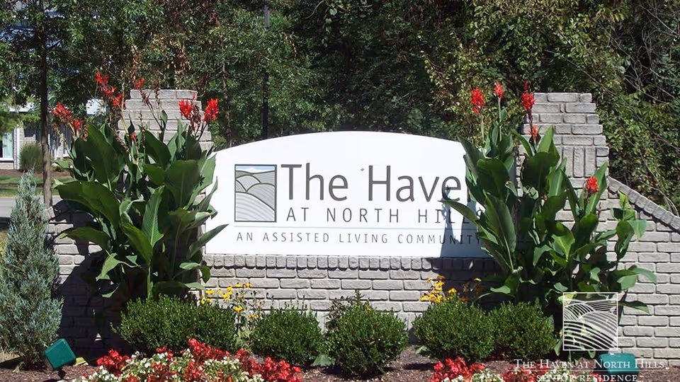 A landscaped entrance sign for The Haven at North Hills, an assisted living community, surrounded by green plants and red flowers with a brick wall backdrop.