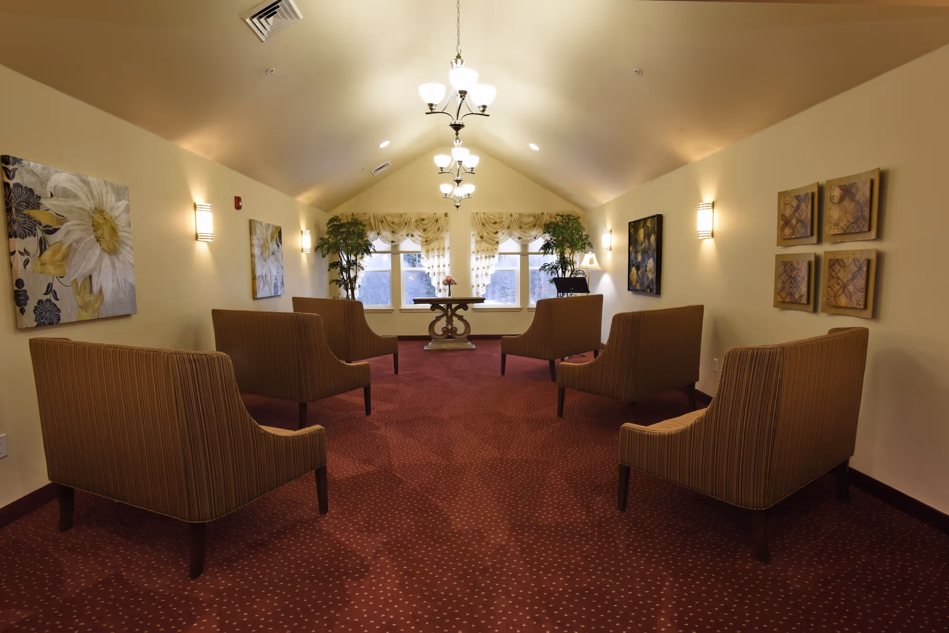 A cozy lounge area with six striped armchairs arranged in two rows facing each other on a red carpet with small patterns. The room has cream-colored walls adorned with floral and abstract paintings, two large potted plants near windows with sheer curtains, and a wooden table with a flower vase in the center. Ceiling lights and wall sconces illuminate the space.