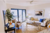 A bright living room with a white sofa adorned with blue and beige cushions, a wooden coffee table with decorative items, two beige armchairs, a side table with a lamp, a potted plant, and large glass doors leading to a balcony with a view of the water.