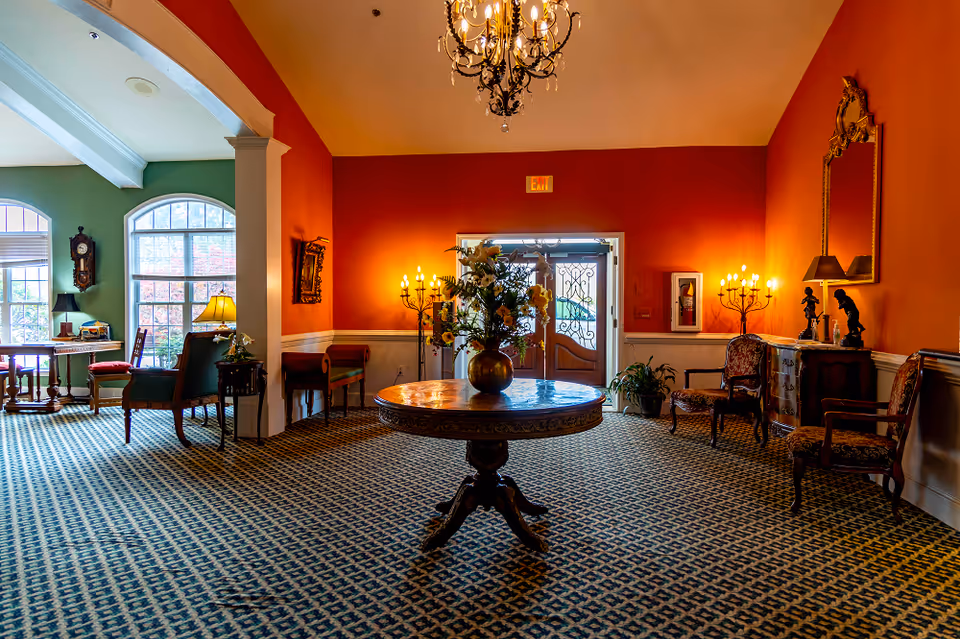 A warmly lit senior living facility interior with a central round wooden table holding a large floral arrangement. The room features patterned carpet, orange and green walls, antique-style chairs, a chandelier, wall sconces, a large mirror, and a decorative clock near large windows letting in natural light.
