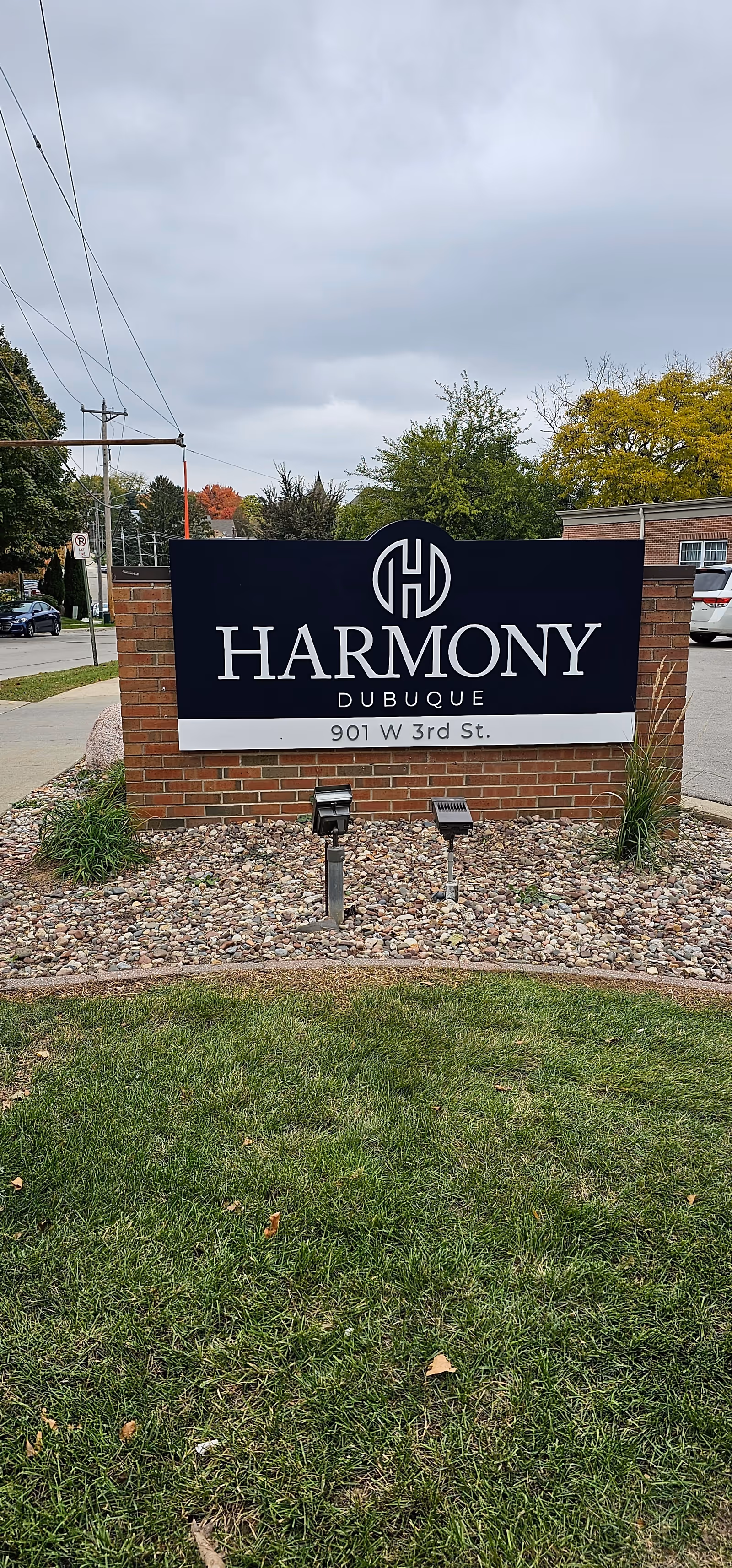 A brick entrance sign reading "Harmony Dubuque 901 W 3rd St." set in a rock bed with grass, trees, and a building in the background.