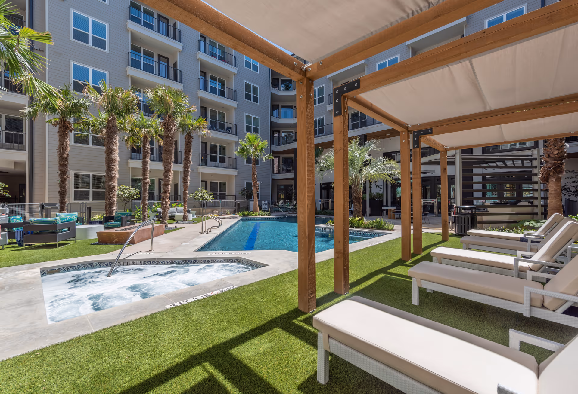 Outdoor pool area at Overture Domain featuring a swimming pool, a hot tub with bubbling water, palm trees, lounge chairs under wooden pergolas with fabric canopies, and a multi-story apartment building in the background.