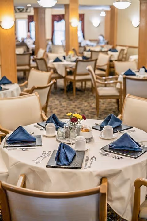 A dining room in a senior living facility with round tables covered in white tablecloths, each set with blue folded napkins, white cups, silverware, and small flower arrangements. The room has beige walls, wooden columns, and soft lighting fixtures.