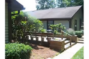 Exterior view of a single-story building with green siding and a gray roof, surrounded by greenery including bushes and trees. There is a wooden ramp with railings leading to the entrance, and a paved walkway beside it.