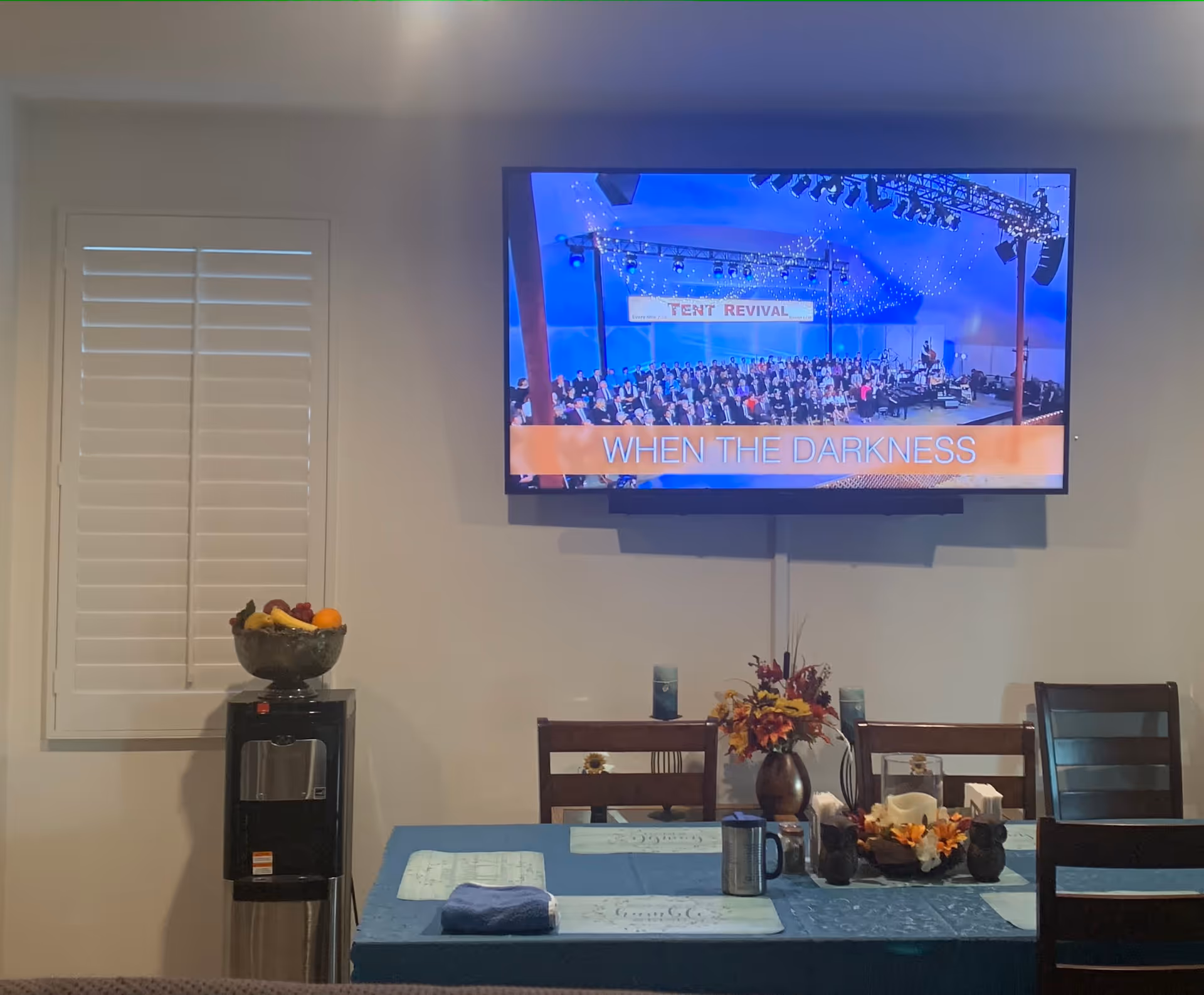 A dining area with a rectangular table covered with a blue tablecloth and placemats. On the table are a vase with artificial flowers, two owl-shaped candle holders, a glass candle holder, a metal travel mug, and a folded towel. Behind the table are wooden chairs. Mounted on the wall above the table is a flat-screen TV displaying a scene with a large group of people and the text 'WHEN THE DARKNESS'. To the left of the table is a water dispenser with a bowl of fruit on top. A window with white shutters is on the left wall.