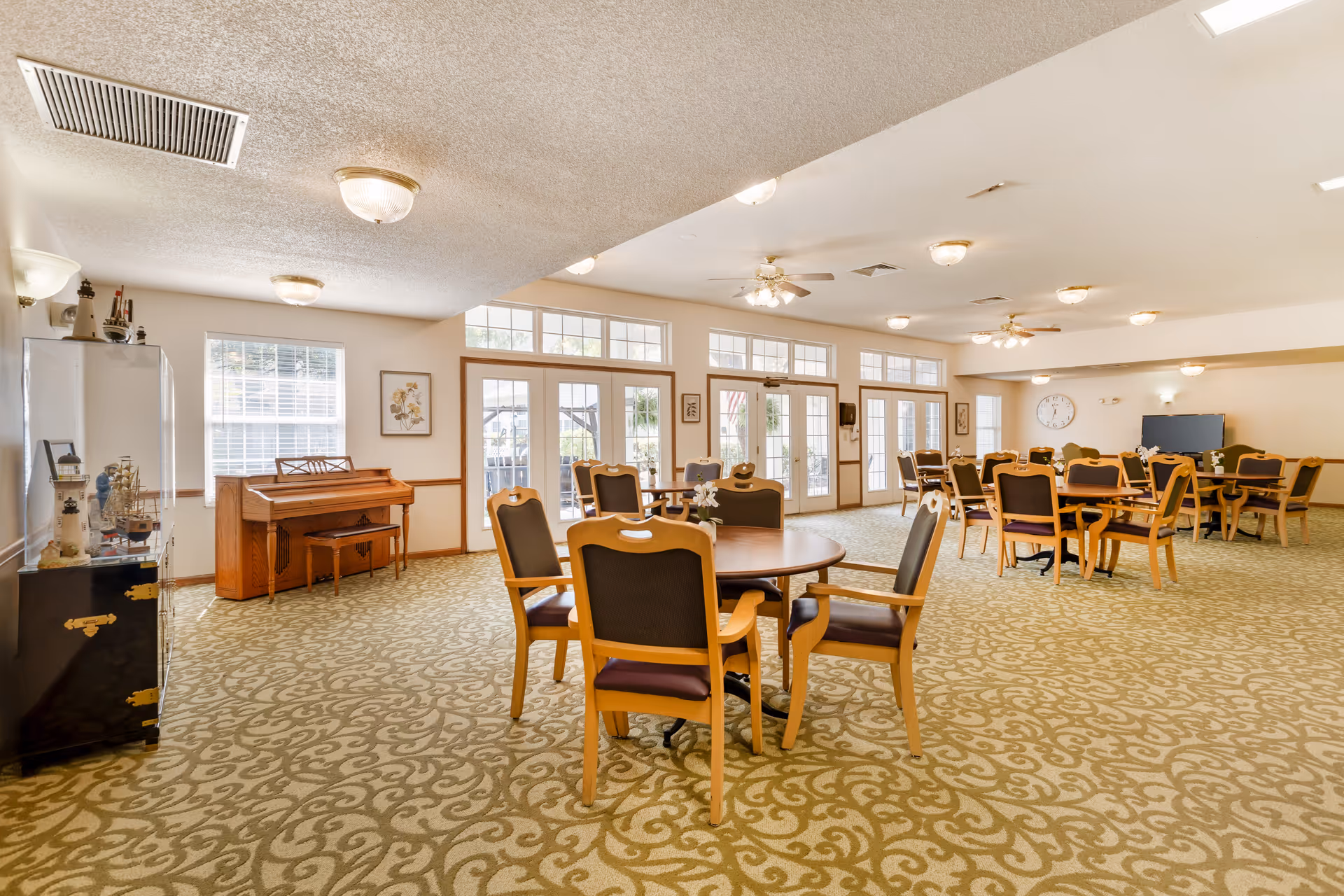 Spacious senior living dining/activity room with multiple round tables and chairs, a piano, and large windows and doors letting in natural light.