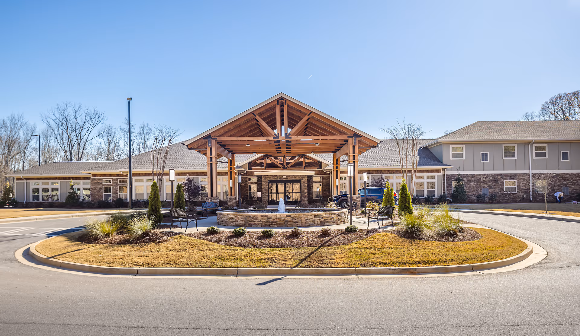 Front exterior view of Oaks at Eagle's Landing facility featuring a circular driveway with landscaped island in the center, benches, a small fountain, and a large wooden entrance canopy with a peaked roof. The building has stone and siding exterior with multiple windows and a clear blue sky in the background.