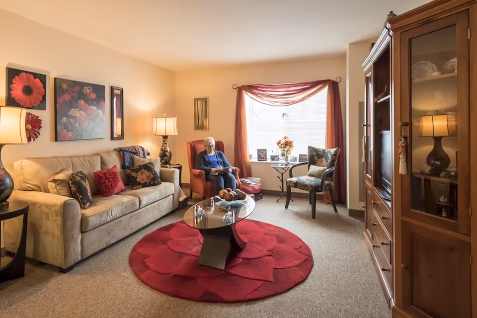 Cozy senior living room with a sofa, armchairs, a glass coffee table on a red round rug, and an elderly woman sitting by a window.