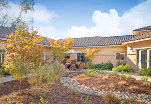Outdoor courtyard area at Cogir of Sonoma Plaza featuring a garden with various plants and small trees, a stone-lined dry creek bed, patio furniture with an umbrella, and a single-story building with a tiled roof under a partly cloudy blue sky.
