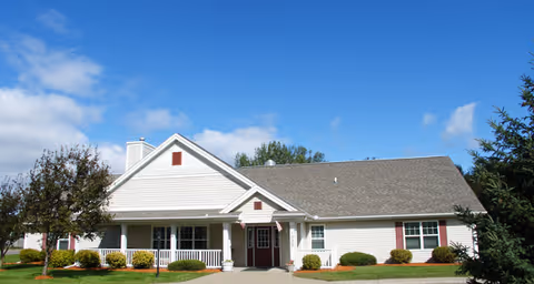 Single-story white residential-style building with a covered front porch, landscaping, and a blue sky background.
