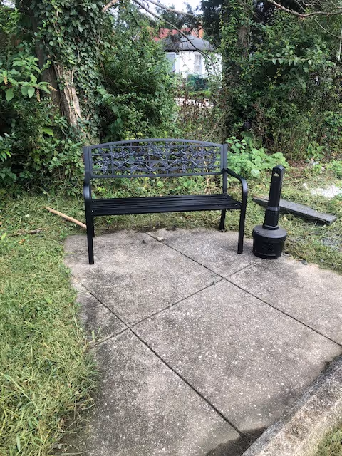 Black metal bench with decorative backrest placed on a concrete patio surrounded by green grass and dense bushes, with a black outdoor heater or lamp next to the bench.