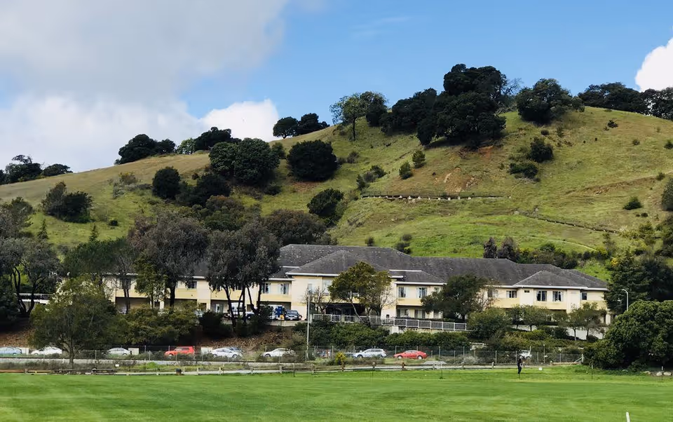 Two-story beige nursing facility building at the base of a tree-covered green hillside with a lawn and parked cars in the foreground.