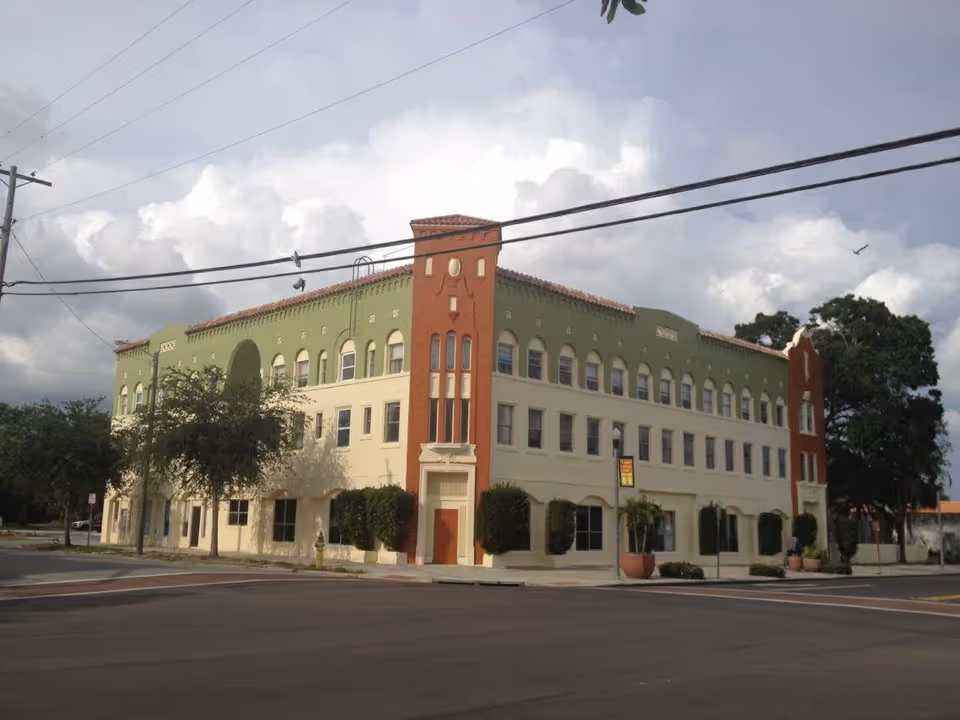 Exterior view of a three-story building with a green and beige facade and red accents on the corners, located at a street intersection under a cloudy sky.