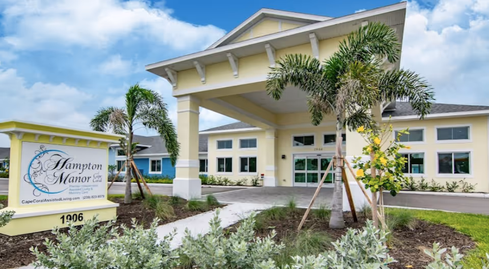 Front exterior view of Hampton Manor of Cape Coral, a single-story assisted living facility with a covered entrance, palm trees, and landscaping under a partly cloudy sky.