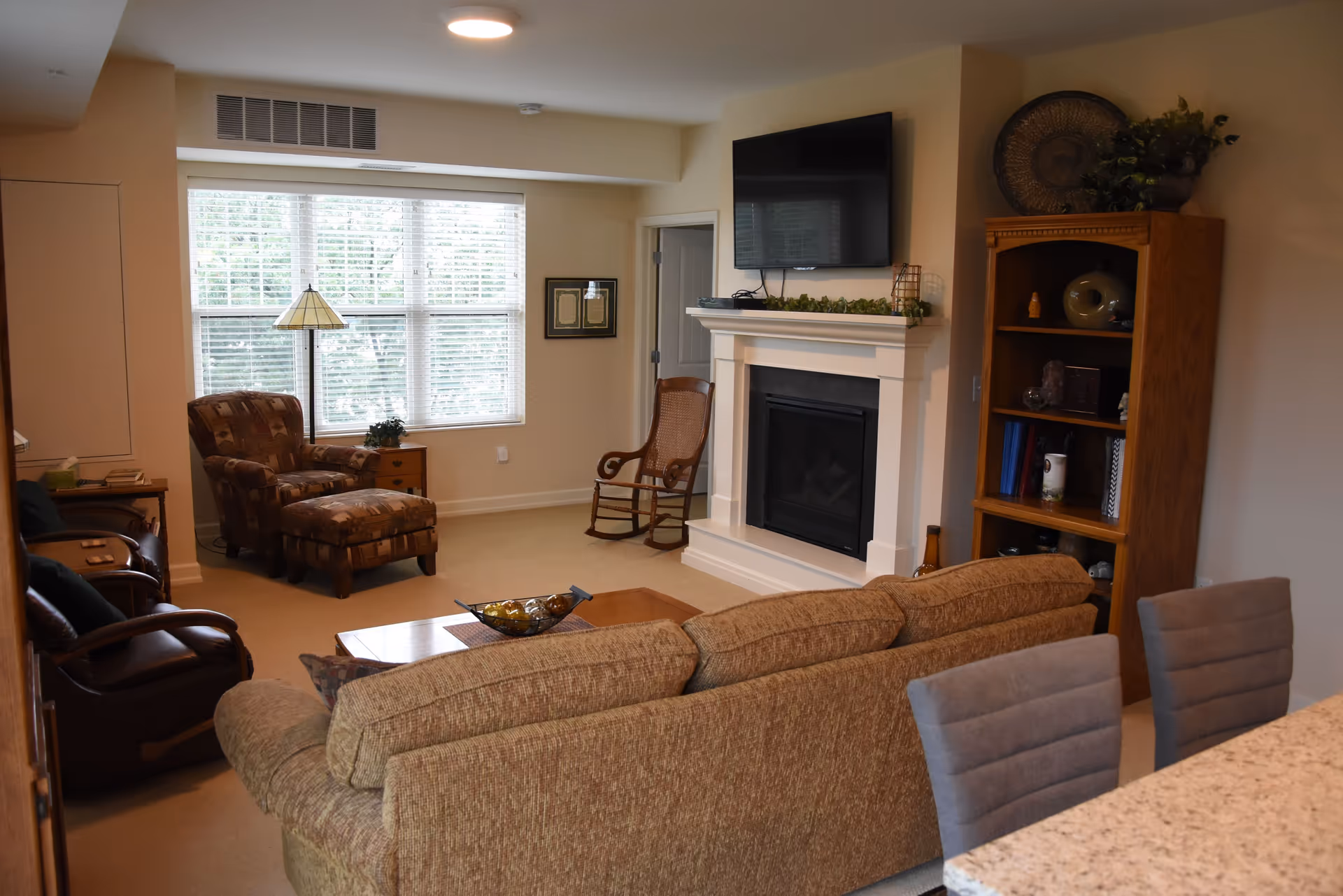 A cozy living room with a beige sofa facing a fireplace topped with a flat-screen TV. There is a wooden rocking chair next to the fireplace, a patterned armchair with an ottoman near a window with white blinds, and a wooden bookshelf with decorative items. Two gray chairs are partially visible at a granite countertop in the foreground.