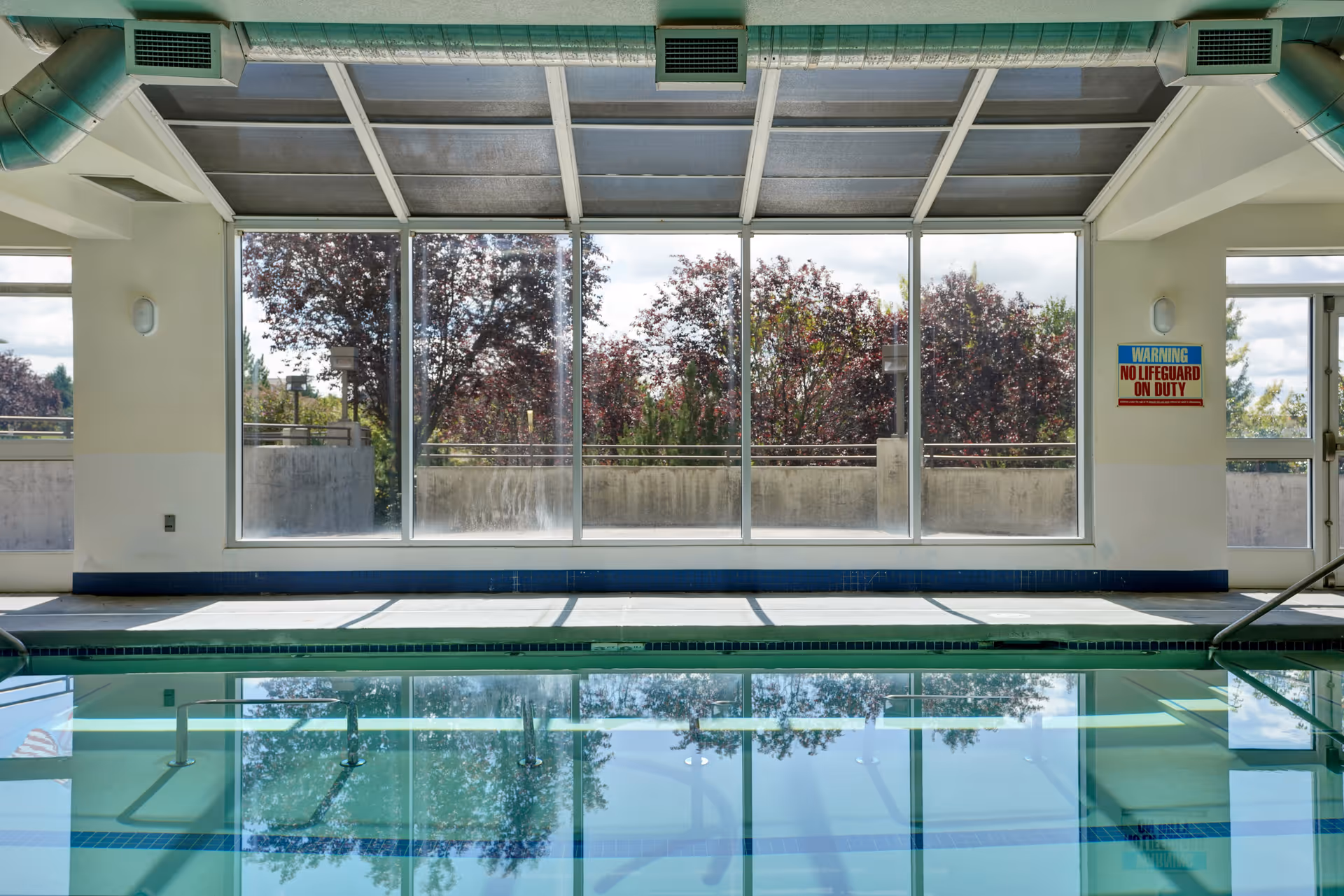 Indoor swimming pool with clear water reflecting large windows that show trees outside. The room has white walls, a blue tiled edge around the pool, and a sign on the wall that reads 'WARNING NO LIFEGUARD ON DUTY'.