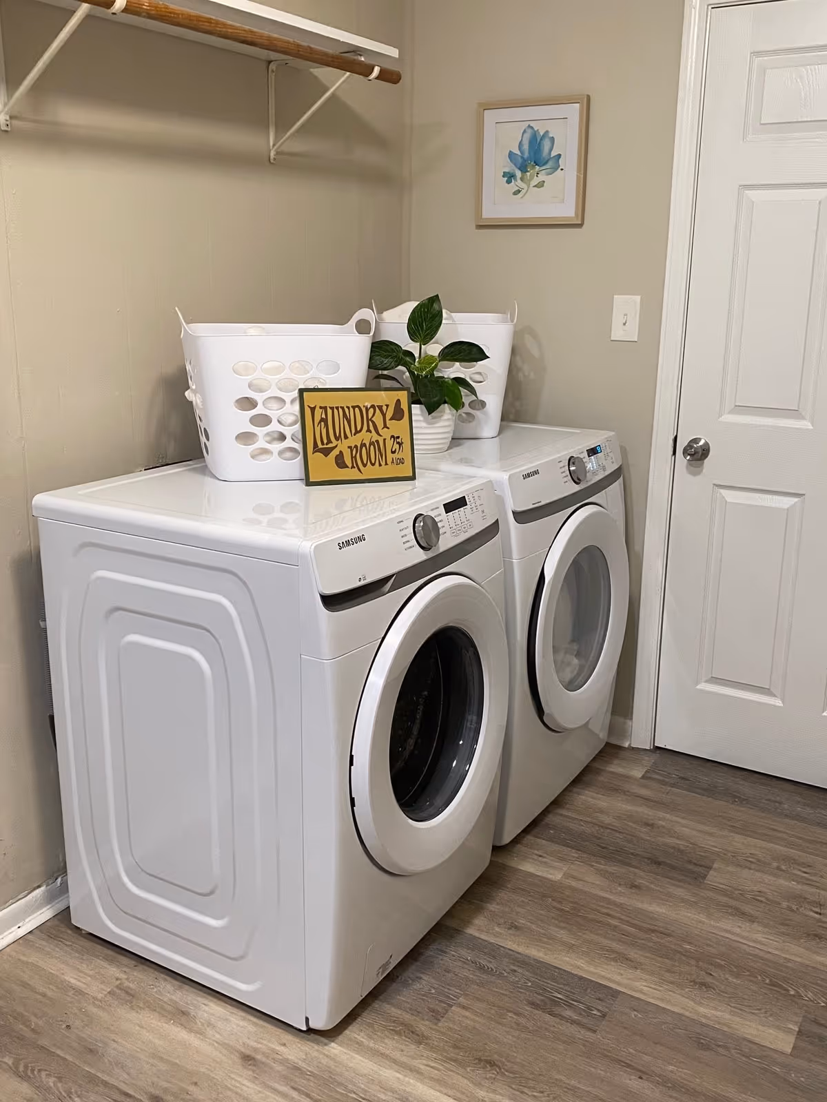Laundry room with a white front-loading washing machine and dryer side by side. On top of the machines are two white laundry baskets, a small green potted plant, and a decorative sign that reads 'Laundry Room 25¢ a load'. A wooden rod for hanging clothes is mounted on the wall above the machines, and a framed picture of a blue flower hangs on the wall next to a closed white door. The floor is covered with wood-patterned vinyl or laminate.