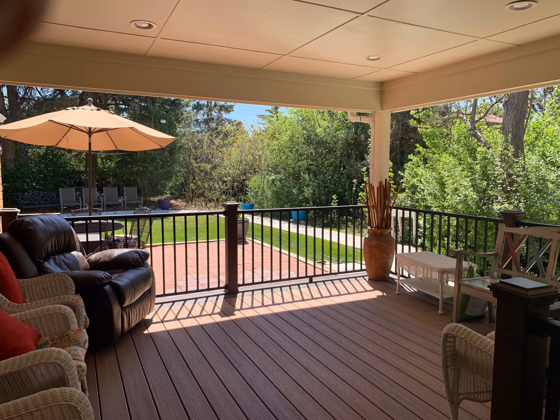 Covered outdoor patio area with wicker and leather seating, a large umbrella shading a table with chairs, and a view of a garden with trees and bushes in the background.