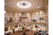 Spacious and elegant interior of a senior living facility with a grand circular staircase in the center, high ceilings with a large circular light fixture, arched windows, and a balcony overlooking the main floor furnished with seating areas and plants.