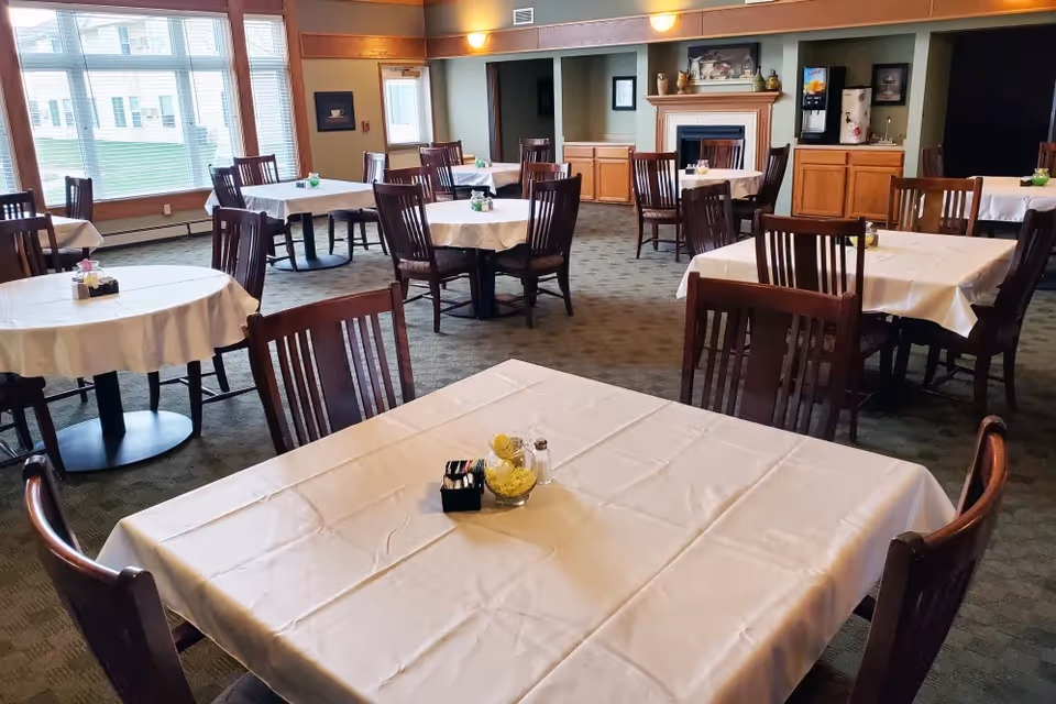 Dining room with tables dressed in white tablecloths and wooden chairs, large windows and a serving area with a fireplace in the background.