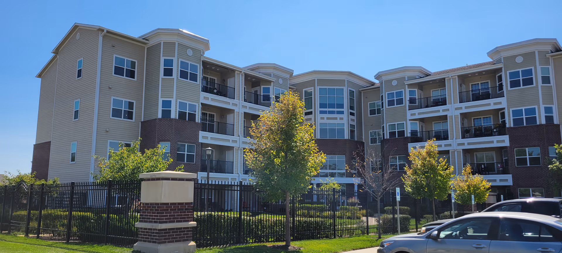 Multi-story senior living apartment building with balconies, a fenced landscaped front yard, trees, and parked cars.