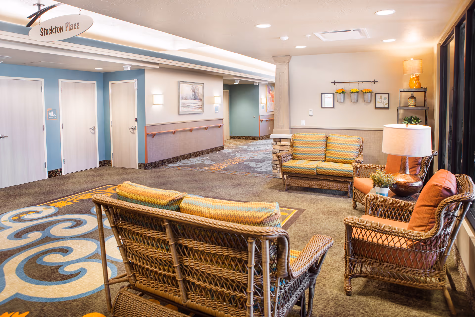 A cozy seating area in a senior living facility hallway with wicker furniture including a loveseat and two armchairs with cushions, a side table with a lamp and plant, and wall decorations with small potted plants. The hallway has multiple doors, a carpet with a decorative pattern, and a sign overhead reading 'Stockton Place'.