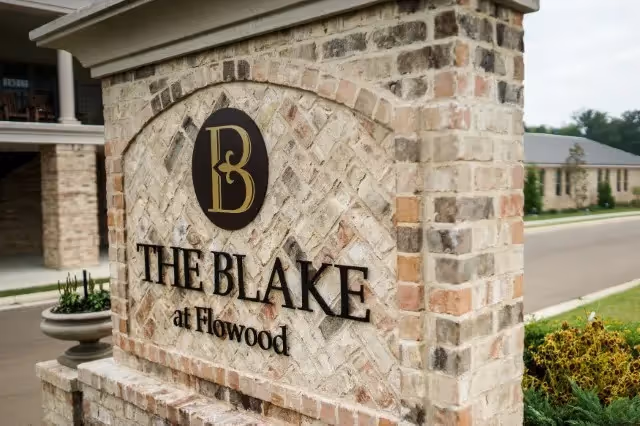 A brick sign at the entrance of a facility displaying the name 'The Blake at Flowood' with a circular logo featuring the letters B and L above the text. The sign is situated near a road with buildings and greenery visible in the background.