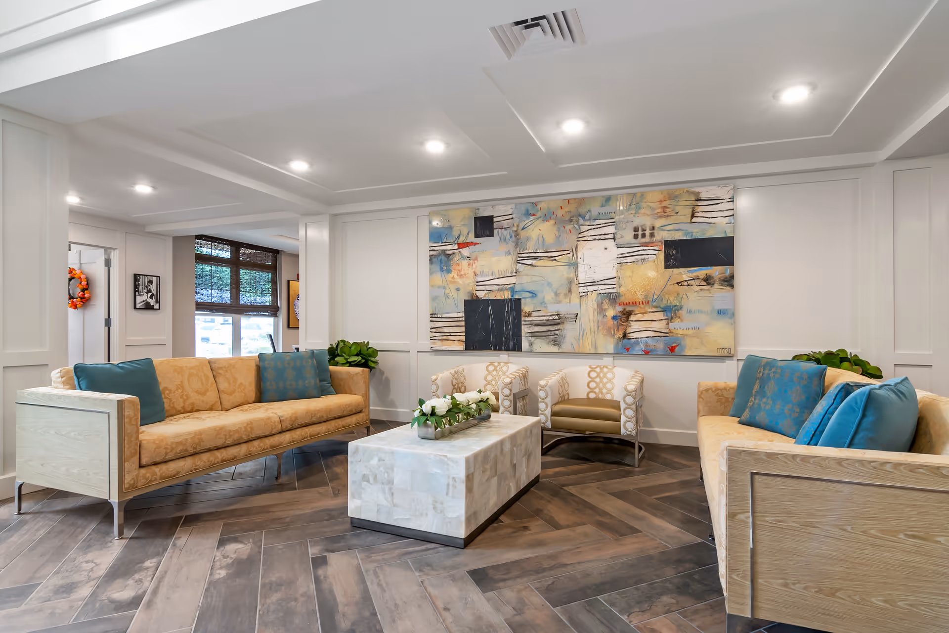 A modern senior living facility lounge area with two beige sofas adorned with blue and patterned cushions, two patterned armchairs, a rectangular marble coffee table with a floral centerpiece, abstract artwork on the wall, and wooden herringbone flooring.