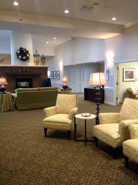 A cozy living room area in a senior living facility featuring patterned beige armchairs, a small round side table with a decorative item, and a tall floor lamp. In the background, there is a green sofa facing a brick fireplace adorned with decorative urns and a wreath. The room has warm lighting, carpeted floors with a geometric pattern, and light-colored walls with framed artwork.