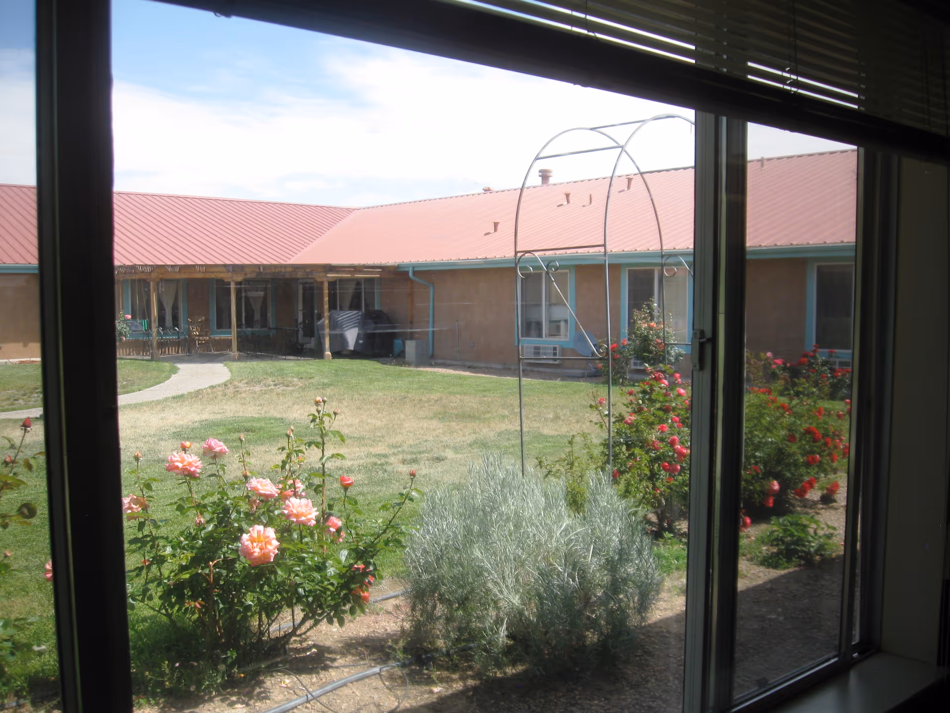 View through a window showing a garden with blooming pink and red roses, green shrubs, and a metal garden arch. In the background, there is a single-story building with a red roof and beige walls.