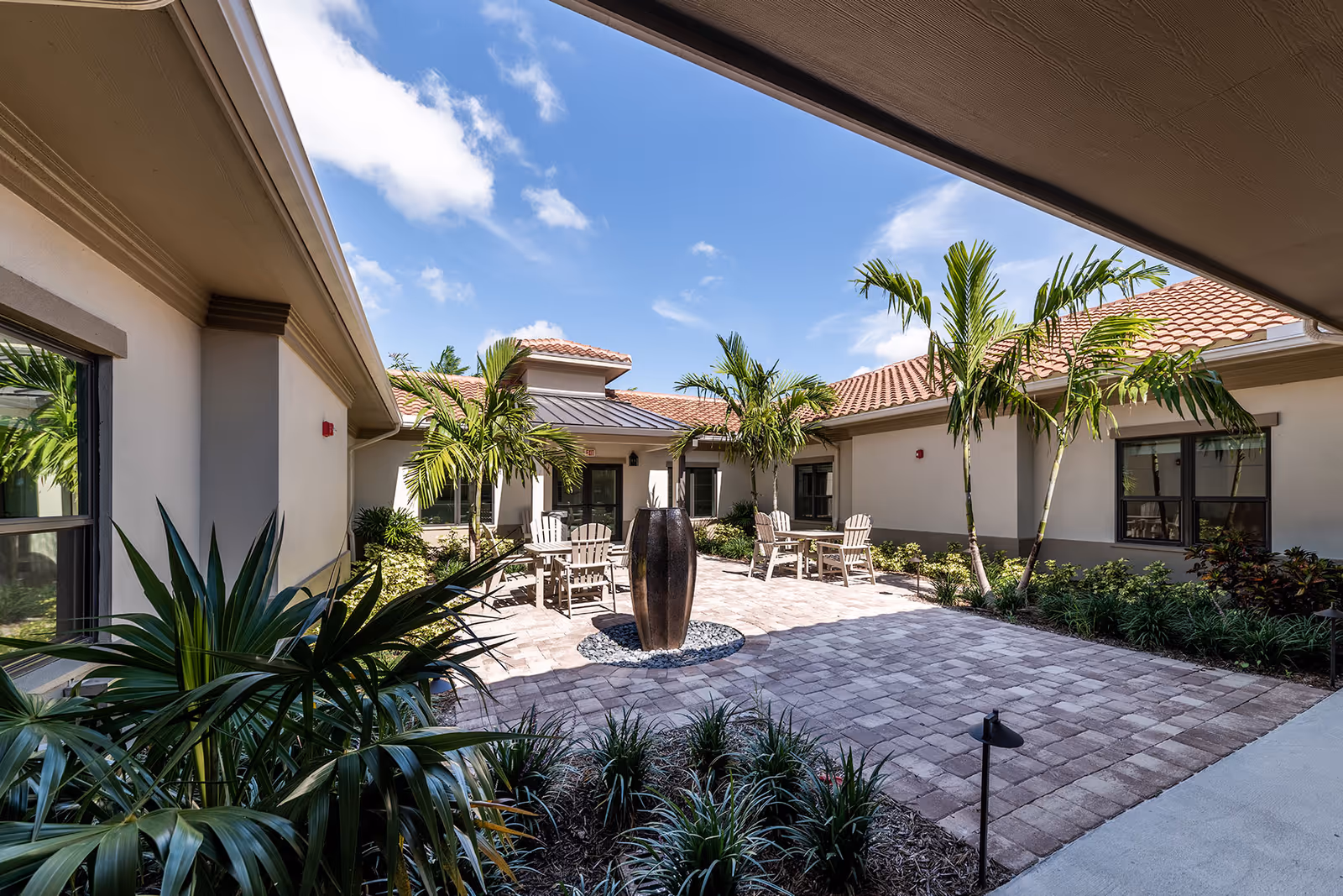 Sunlit courtyard with a central water feature, Adirondack chairs, palm trees and surrounding single-story buildings.