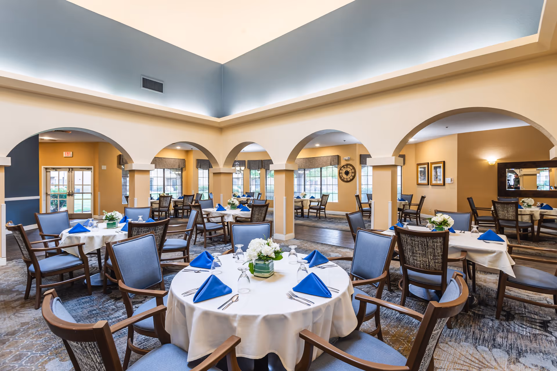 A bright and spacious dining room with round tables covered in white tablecloths, each set with blue folded napkins, glassware, and silverware. The room features arched doorways, large windows letting in natural light, and comfortable wooden chairs with blue cushions. Floral centerpieces decorate the tables, and the walls are painted in soft beige and blue tones.