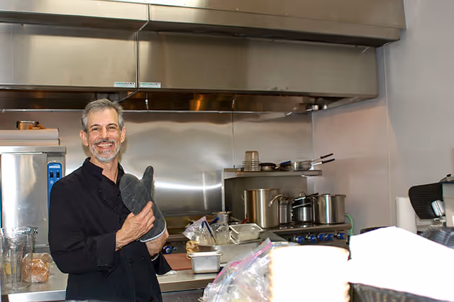 Smiling chef holding an oven mitt in a stainless-steel commercial kitchen with pots and pans.