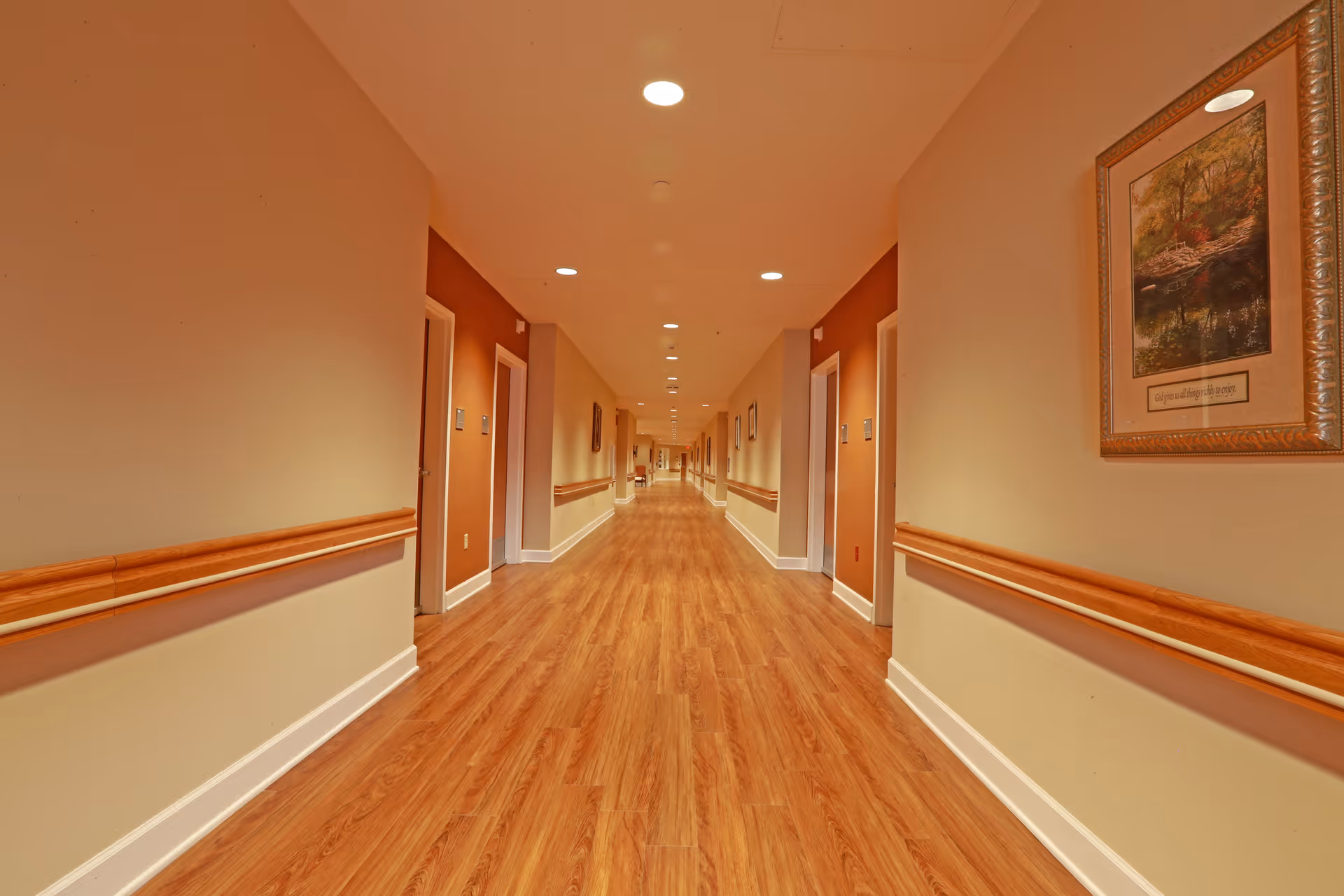 Long, well-lit interior hallway with wood flooring, handrails, recessed ceiling lights and doors on both sides.