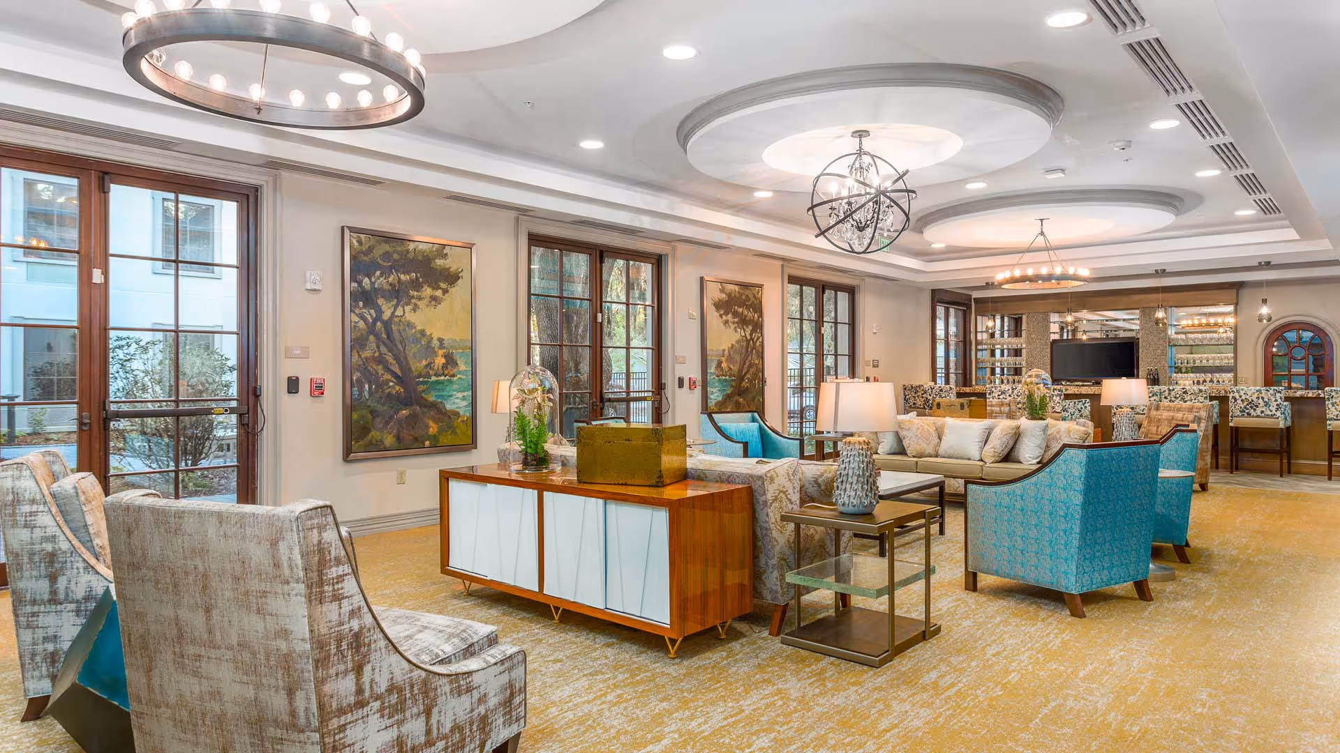 A spacious and elegantly furnished living room area with multiple seating options including patterned armchairs and blue upholstered chairs arranged around a central wooden cabinet. The room features large windows with wooden frames, allowing natural light to enter. The ceiling has decorative circular recesses with modern chandeliers. There are landscape paintings on the walls and a bar area with stools in the background.