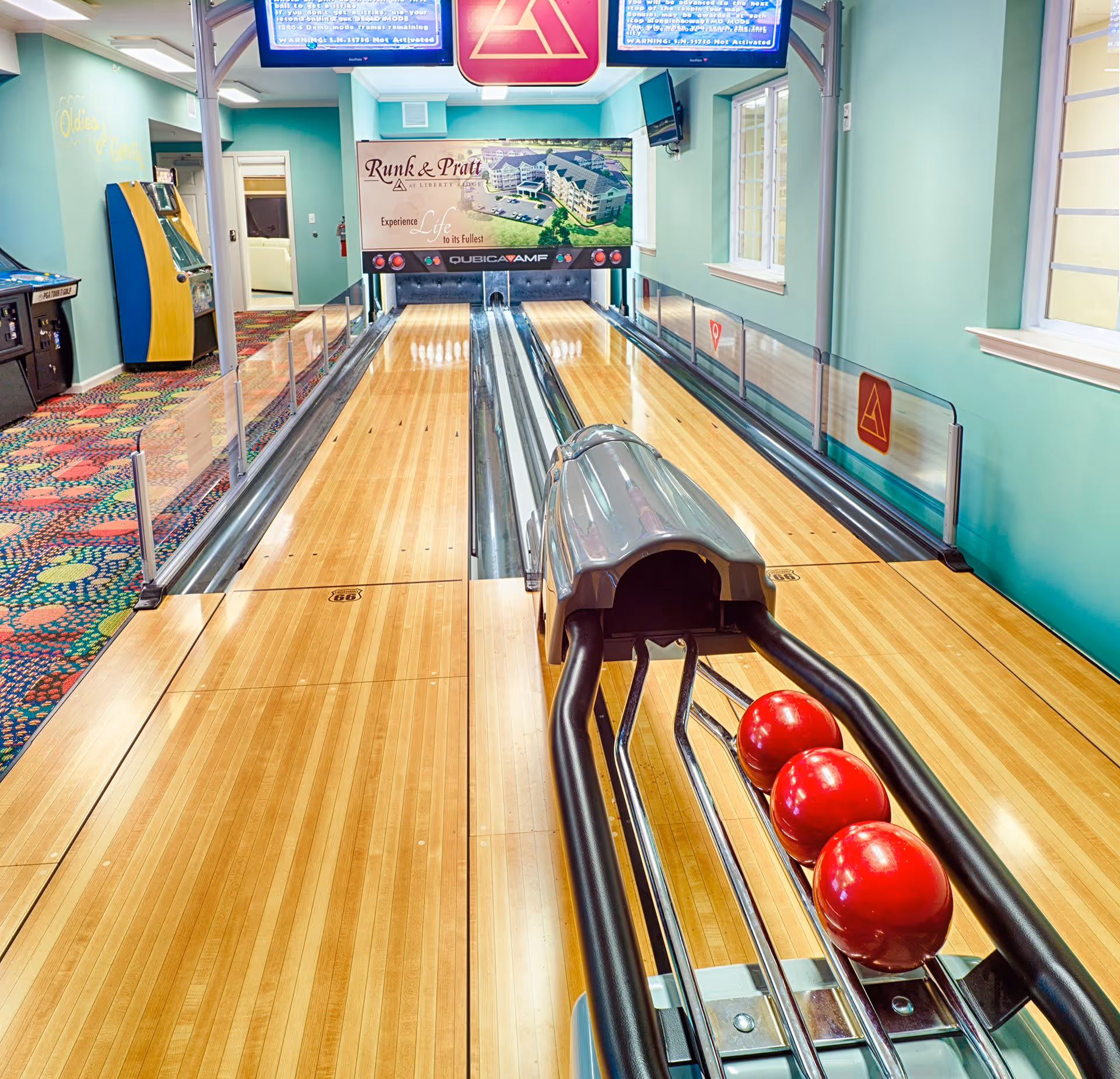 Indoor bowling alley with two lanes, three red bowling balls on the ball return, colorful patterned carpet on the left side, and a sign at the end of the lanes that reads 'Runk & Pratt at Liberty Ridge Experience Life to its Fullest'.