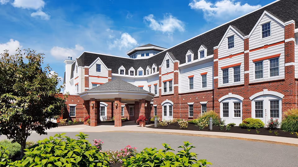 Exterior view of a large senior living facility building with red brick and white siding, multiple windows, and a covered entrance. The foreground features a circular driveway, landscaped bushes, flowers, and a tree under a blue sky with some clouds.