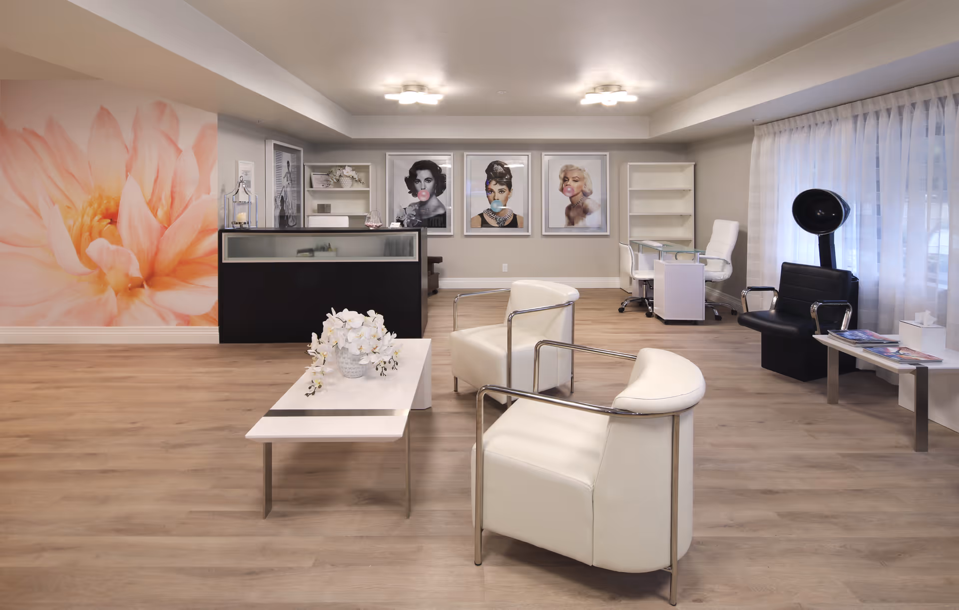 A bright modern lounge/reception area with white armchairs, a coffee table with flowers, a reception desk, a large floral wall mural and framed portraits.
