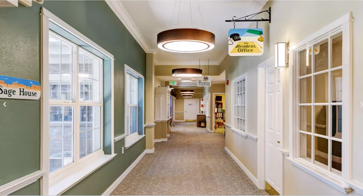 A well-lit hallway in a senior living facility with green and beige walls, large windows on the left, and doors and windows on the right. Hanging signs indicate directions to the Resident Office and the Whispering Fellowship. Modern circular ceiling lights illuminate the carpeted corridor.