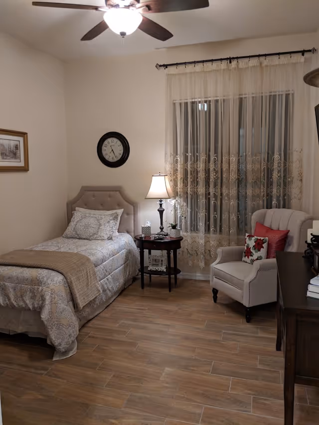 A cozy bedroom with a single bed covered in patterned bedding and a beige throw blanket. Next to the bed is a round wooden side table with a lamp, a small plant, and a tissue box. A beige armchair with red and floral pillows is placed near a window with sheer lace curtains. The room has wooden flooring, a ceiling fan with a light, a wall clock, and a framed picture on the wall.