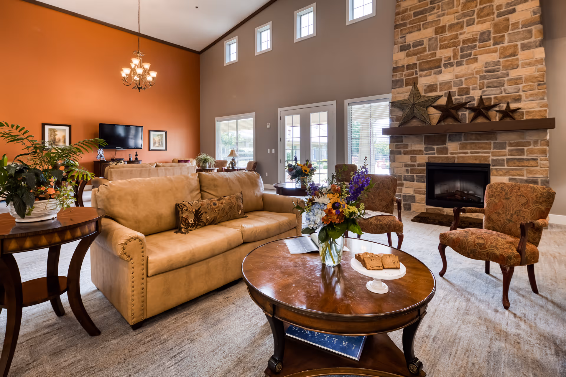Bright communal living room featuring a tan sofa, patterned armchairs, a round coffee table with flowers and snacks, and a stone fireplace.