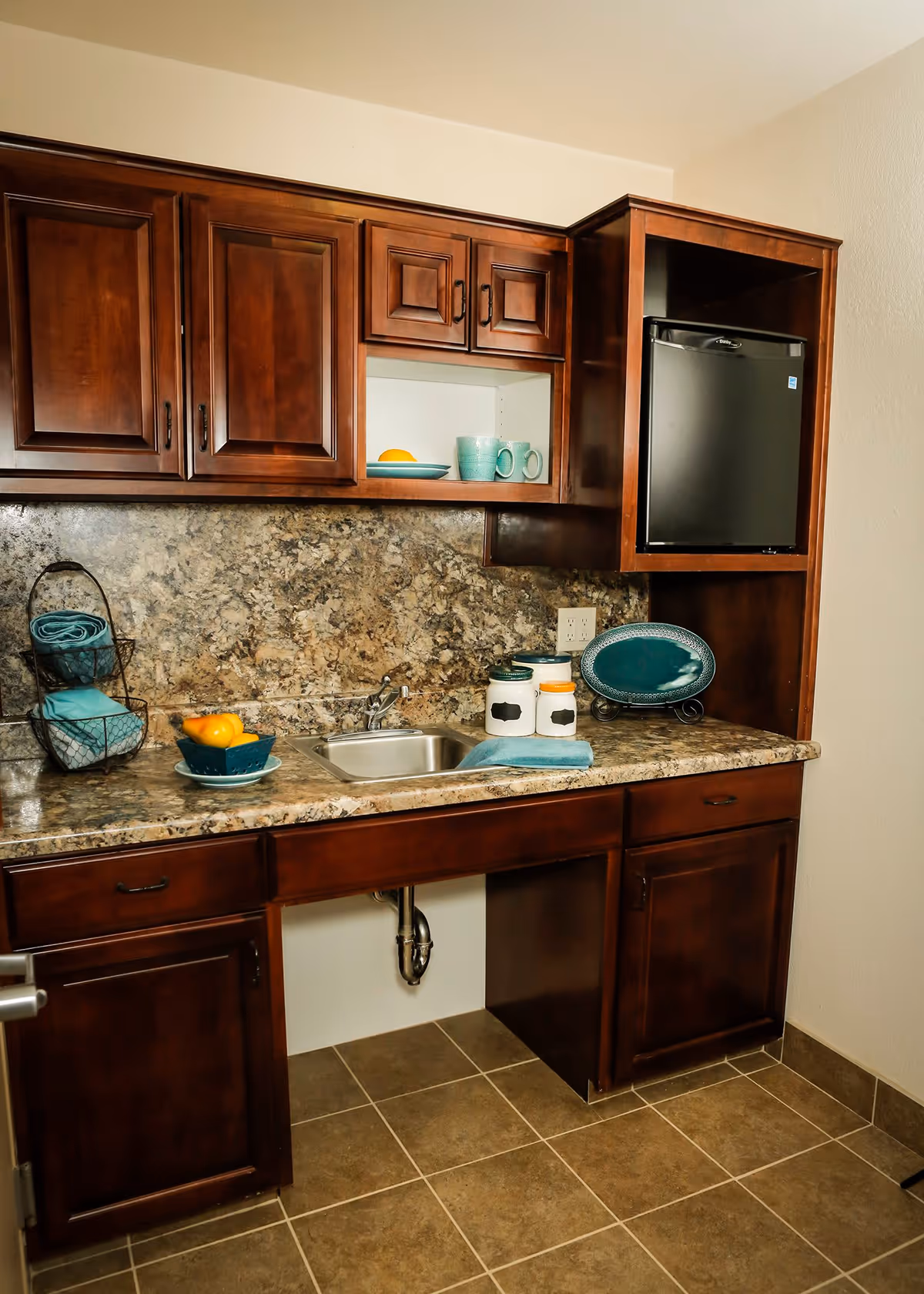 A kitchenette area with dark wooden cabinets, a small stainless steel sink, a mini refrigerator, and a granite countertop. On the counter are decorative items including a bowl of oranges, two ceramic canisters, a teal towel, and a teal plate on a stand. The backsplash matches the granite countertop, and the floor is tiled in a brown tone.