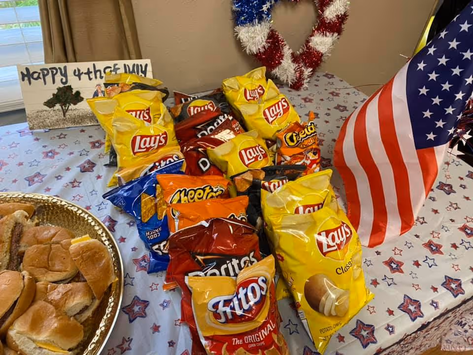 A table decorated with a patriotic theme featuring an American flag, a red, white, and blue wreath, and a sign that says 'Happy 4th of July'. On the table are multiple bags of chips including Lay's, Doritos, Cheetos, Fritos, and a plate with several sandwiches.