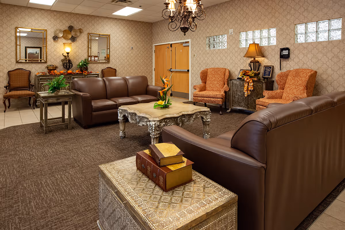 A cozy living room area with two brown leather sofas facing an ornate coffee table with a floral centerpiece. The room features patterned wallpaper, two orange upholstered armchairs, a side table with a lamp, decorative mirrors, and additional seating chairs along the wall. The floor is carpeted with a brown rug, and there are double wooden doors in the background.
