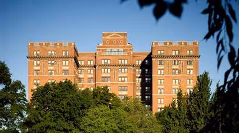 Front-facing brick senior living building rises above green trees against a blue sky.