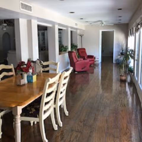 A long room with wooden flooring featuring a wooden dining table with six chairs on the left side and two red recliner chairs further back. There are several windows on the right side letting in natural light, and some potted plants are placed along the room. The walls are light-colored, and there is a ceiling fan and recessed lighting.