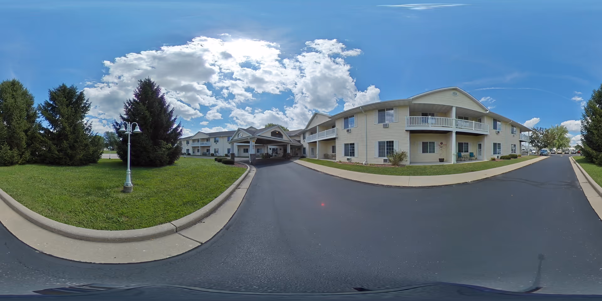 Exterior view of Charleston House Assisted Living facility on a sunny day with a partly cloudy sky. The building is two stories with balconies and a covered entrance. There is a paved driveway and well-maintained green lawn with trees and a lamp post.